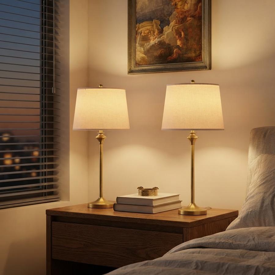 Brass gold table lamp lit beside a bed, hand at the socket